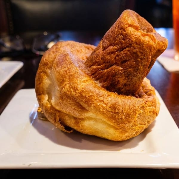 Large golden-brown Yorkshire pudding on a white plate, with a drink and another plate in the background.